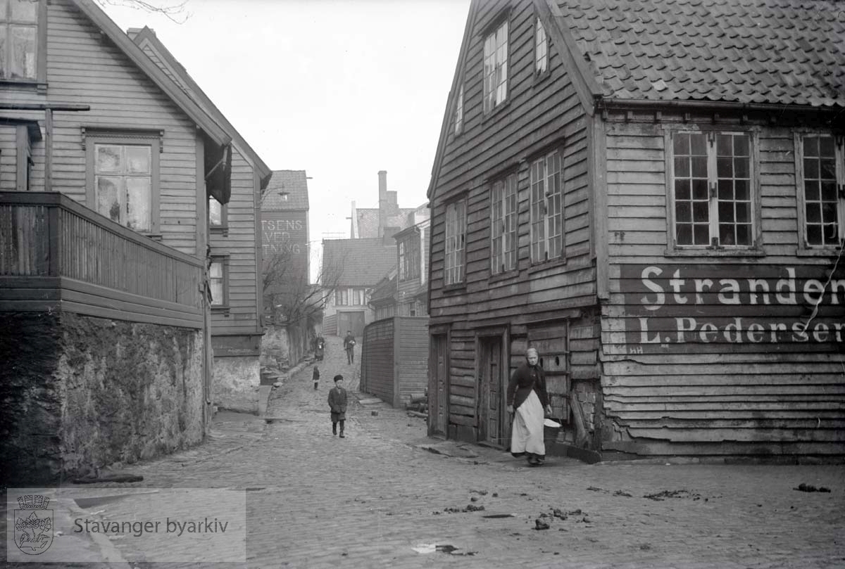 Til høyre, Strandens Kullforretning, N. Strandgt. 53. Til venstre, N. Strandgt. 50 og 52. Mellom den går Andasmauet opp. I bakgrunnen til venstre, Sivertsens Kull- og Vedforretning, N. Strandgt. 60. Bak til høyre, N. Strandgt. 63 og 59.