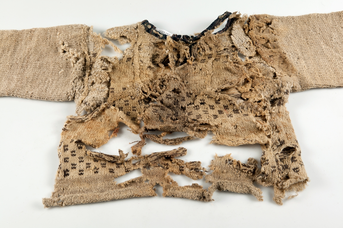 Fragment av stickad tröja med mönsterstickad bård i brunt och beige ullgarn. Bålen i 1-trådigt ullgarn mönsterstickad i rutmönster i brunt, natursvart och beige. Ärmarna är rundstickade i 2-trådigt ullgarn i naturvit, natursvart, brunt och beige. Sprund mitt fram 10 cm långt. Halsurringning och sprund mitt fram kantad med samma tyg som finns som lagningar på UM23252 och A 38 (UM ?). Hyska och hake i mässing mitt fram.