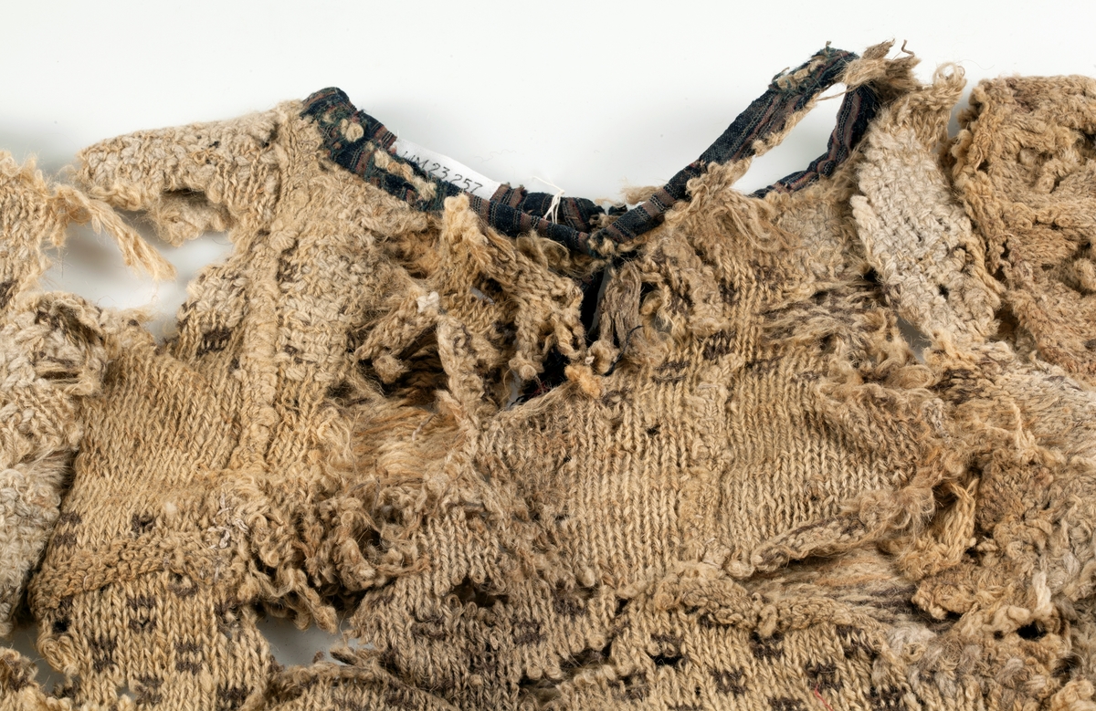 Fragment av stickad tröja med mönsterstickad bård i brunt och beige ullgarn. Bålen i 1-trådigt ullgarn mönsterstickad i rutmönster i brunt, natursvart och beige. Ärmarna är rundstickade i 2-trådigt ullgarn i naturvit, natursvart, brunt och beige. Sprund mitt fram 10 cm långt. Halsurringning och sprund mitt fram kantad med samma tyg som finns som lagningar på UM23252 och A 38 (UM ?). Hyska och hake i mässing mitt fram.