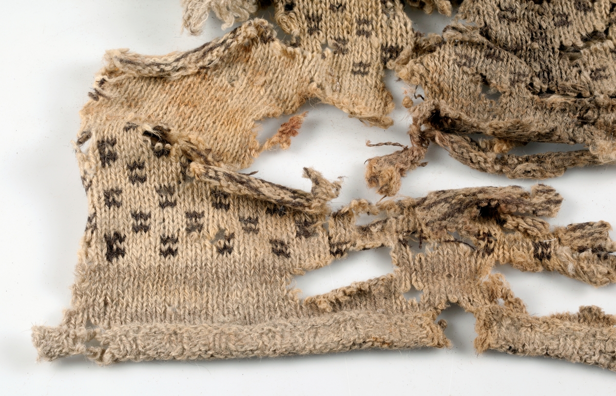 Fragment av stickad tröja med mönsterstickad bård i brunt och beige ullgarn. Bålen i 1-trådigt ullgarn mönsterstickad i rutmönster i brunt, natursvart och beige. Ärmarna är rundstickade i 2-trådigt ullgarn i naturvit, natursvart, brunt och beige. Sprund mitt fram 10 cm långt. Halsurringning och sprund mitt fram kantad med samma tyg som finns som lagningar på UM23252 och A 38 (UM ?). Hyska och hake i mässing mitt fram.