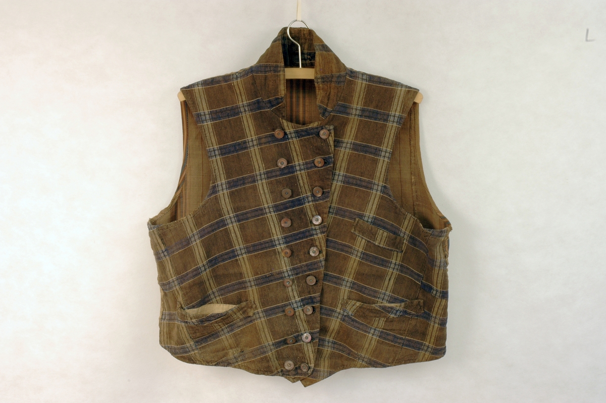 Dobbeltspent vest i rutete stoff i halvull. Lukking i front med ni perlemorsknapper/knapphull. Brystlomme venstre side, og stolpelommer på begge sider. Liten krage. Linfór i front. Stripete bomullstoff i ryggen.