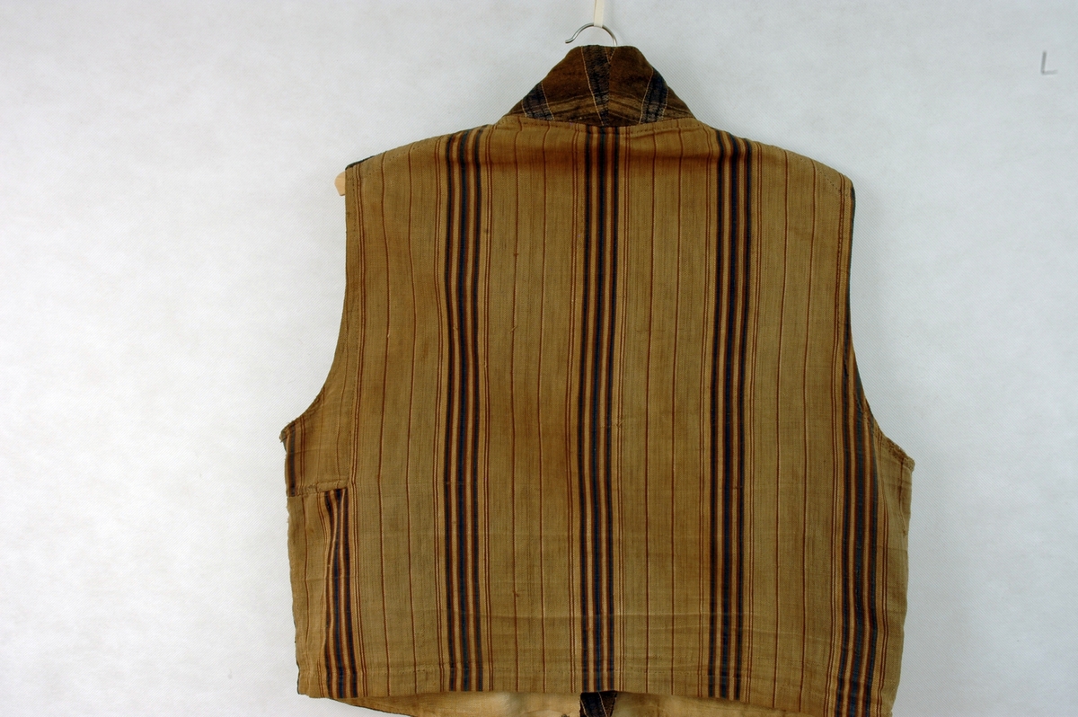 Dobbeltspent vest i rutete stoff i halvull. Lukking i front med ni perlemorsknapper/knapphull. Brystlomme venstre side, og stolpelommer på begge sider. Liten krage. Linfór i front. Stripete bomullstoff i ryggen.