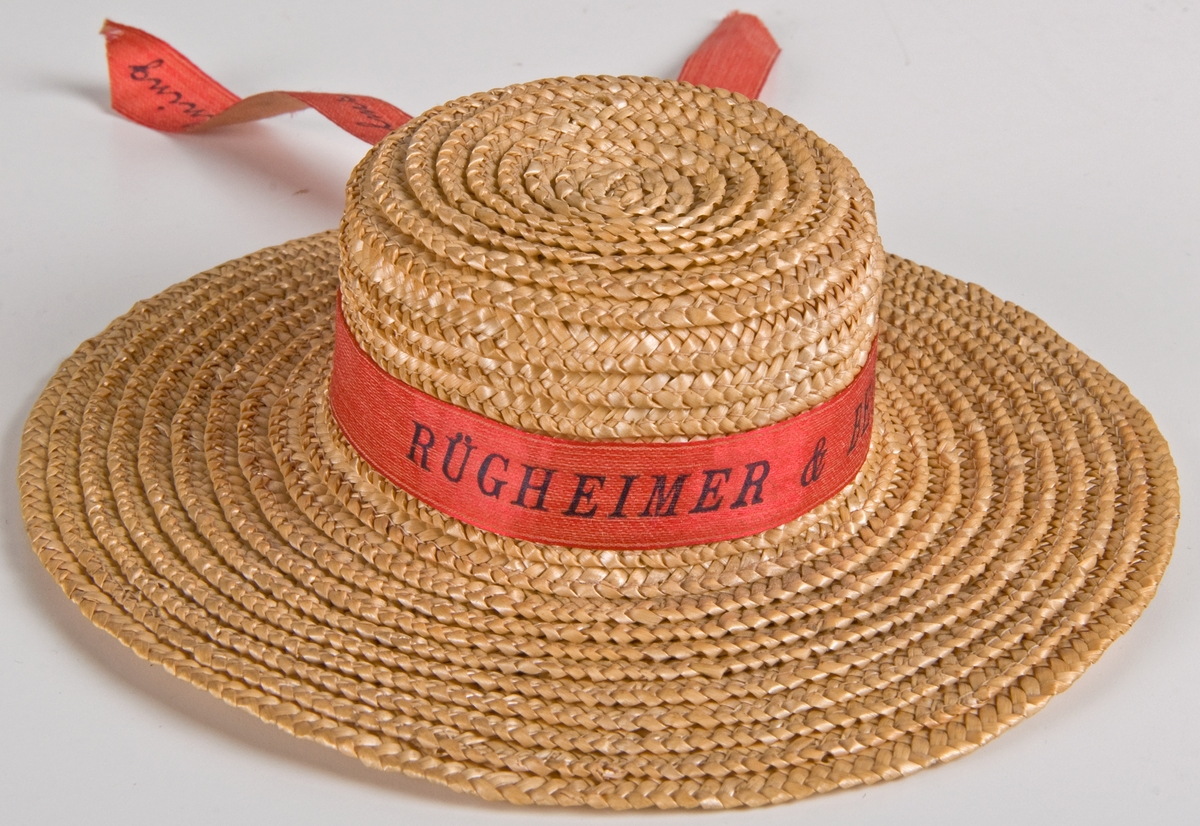 Två dockhalmhattar med rött respektive blått band.
a) Med flätat kant och blått band med med guldtexten: "RÜGHEIMER & BECKER, 1897, Minne af Stockholms utställning." Diam 14,5cm.
b) Med rött band och svart text: som a. Diam 14,5.