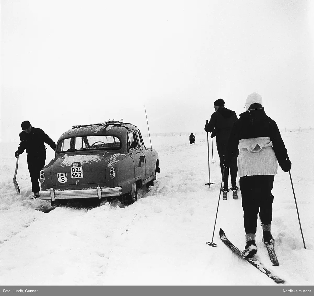 En man med skyffel står vid en Standard Vanguard som kört fast, Södermanland år 1956. Tre personer på skidor åker förbi. Marken är snötäckt.