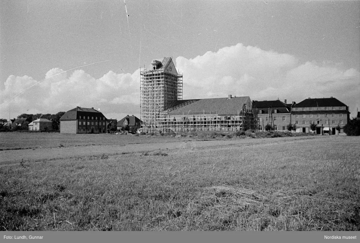 Exteriör av Höganäs kyrka, Himmelfärdskyrkan, under byggnation och som nybyggd, en kyrkobyggnad omgiven av byggnadsställningar, två bilar parkerade framför en kyrka.