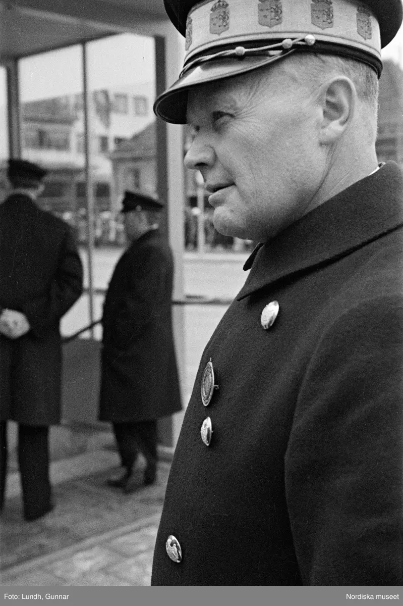 Invigning av Södra Bantorgets (Medborgarplatsen) tunnelbanestation på Södertunneln den 30 september år 1933, Kung Gustaf V kliver in i en bil, porträtt av en man i uniform - en chaufför vid Kungliga Hovstallet, detalj av en bil med lykta och skärm, en grupp män går ut ur en tunnelbanenedgång på Södra Bantorget - män i uniform gör honnör och i bakgrunden står människor i öppna fönster.