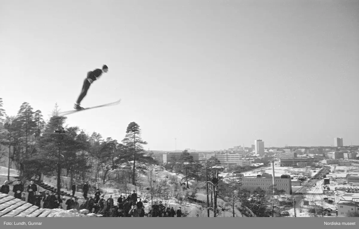 En tävling i backhoppning i Hammabybacken, Stockholm, backhoppare hoppar från en hoppbacke inför åskådare och domare, vy över Södra Hammabyhamnen och Södermalm.