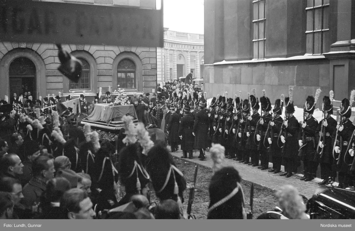 Troligen begravningen av Prins Gustaf Adolf, en kista bärs ut från Storkyrkan, kungafamiljen går ut från Storkyrkan, militärer i uniform står i givakt, kistan står på en katafalk som dras av hästar, en folksamling står längs vägen när begravningsföljet passerar.