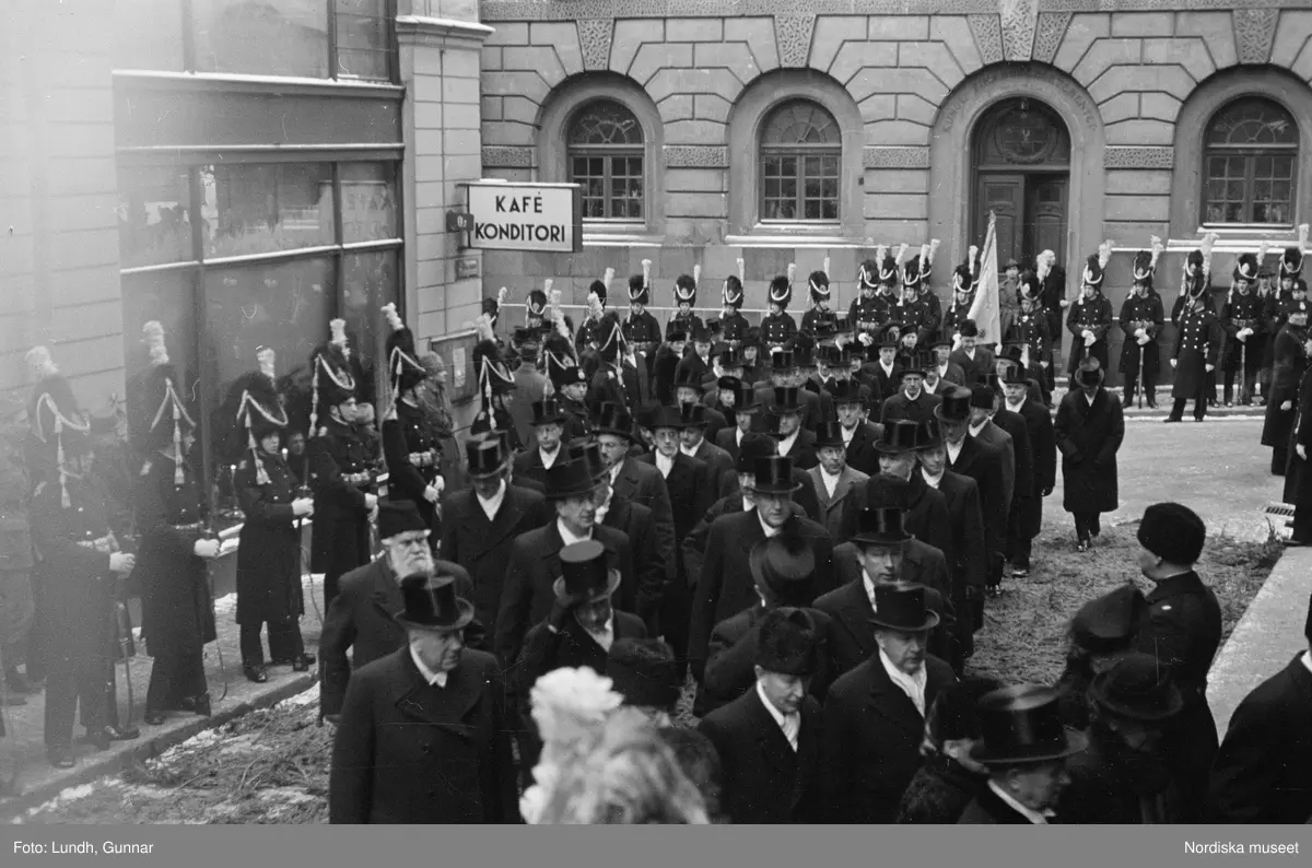 Troligen begravningen av Prins Gustaf Adolf, en kista bärs ut från Storkyrkan, kungafamiljen går ut från Storkyrkan, militärer i uniform står i givakt, kistan står på en katafalk som dras av hästar, en folksamling står längs vägen när begravningsföljet passerar.