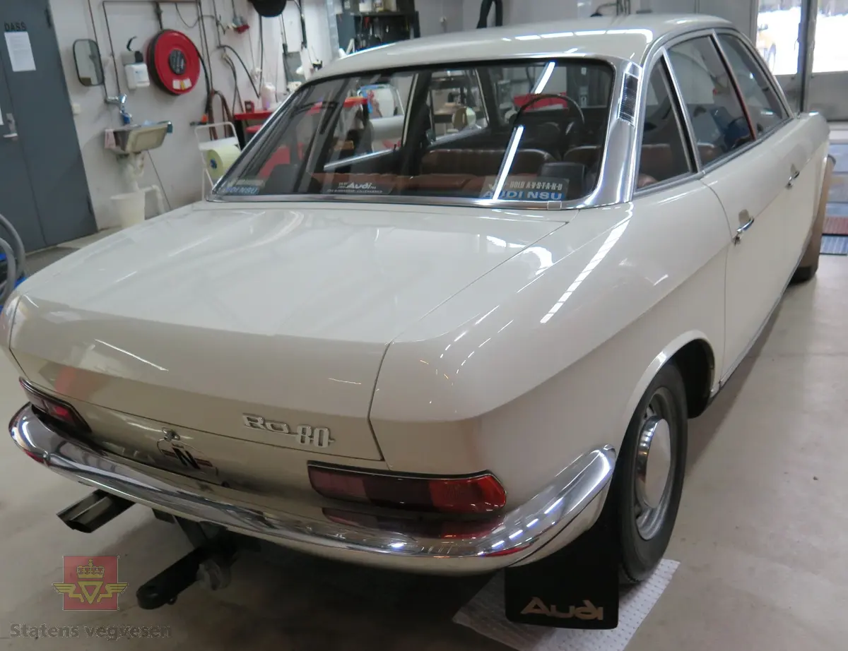 NSU RO 80. 4-dørs hvit bil med brunt interiør. Bilen har en bensindrevet dobbel NSU/Wankel rotasjonsstempelmotor, med sylindervolum på 994 kubikkcentimeter (cm3), og effekt på 115 DIN HK. Dekkdimensjon foran og bak skal standard være 175 HR 14. Framhjulstrekk. Girkasse med 3 gir framover pluss revers. Kilometerstand er 75385.