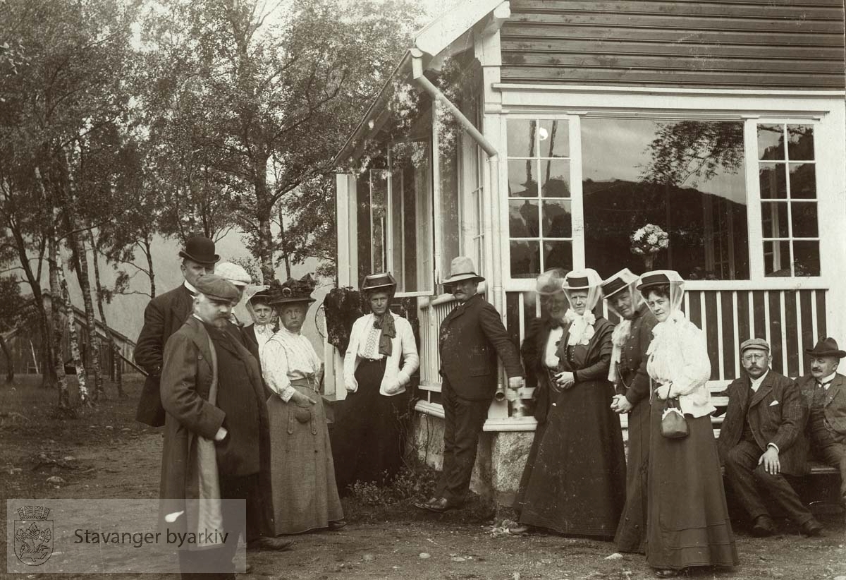 Eiendommen tilhørte statsråd C. Middelthon. En glimrende lakseelv i nærheten. .Fra venstre: Karl G. Kielland, Dagfin H. Berner (hvit caps), Erik Berentsen (sort hatt), fru Rachel Bergesen (delvis skjult), fru Olga Middelthon, Olga Berentsen, Cornelius Middelthon, Lina Gabrielsen, fru Robertsen Larsen (?), fru Martha Monsen, fru Anna Kielland, Jonas Schanche Monsen (sittende på benk) og Andreas Robertsen Larsen..Bildet er medtatt i hytteboken fra Helle..(Etter opplysninger gitt av Trygve Middelthon)