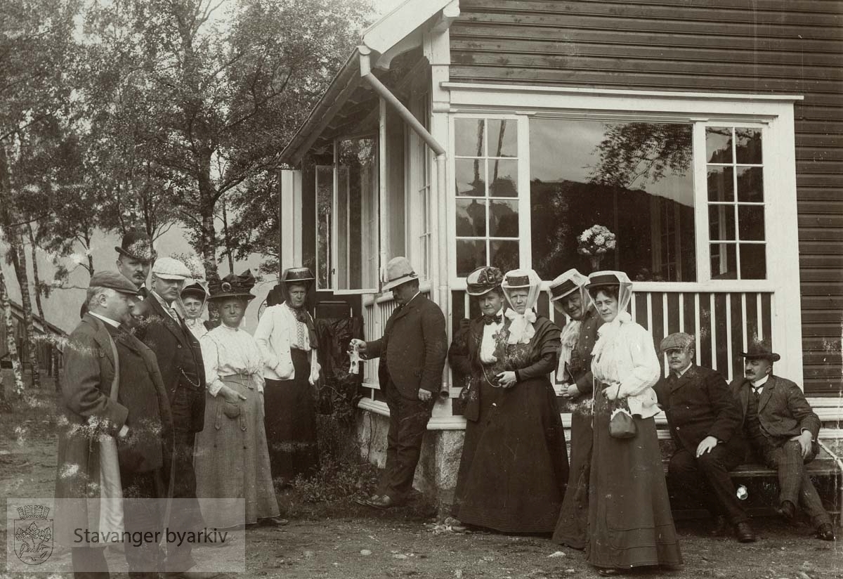 Eiendommen tilhørte statsråd C. Middelthon. En glimrende lakseelv i nærheten. .Fra venstre: Karl G. Kielland, Dagfin H. Berner (hvit caps), Erik Berentsen (sort hatt), fru Rachel Bergesen (delvis skjult), fru Olga Middelthon, Olga Berentsen, Cornelius Middelthon, Lina Gabrielsen, fru Robertsen Larsen (?), fru Martha Monsen, fru Anna Kielland, Georg T. Monsen (sittende på benk) og Andreas Robertsen Larsen..Bildet er medtatt i hytteboken fra Helle..(Etter opplysninger gitt av Trygve Middelthon)