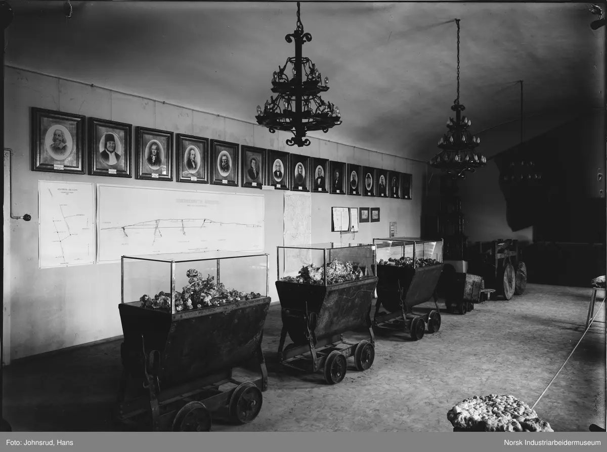 Fylkesutstillingen Kongsberg 1924. Utstilling av vagger med innhold som er glasset inn, antatt sølv fra sølvverket. Lysekroner i taket og bilder av menn på veggen i bakgrunnen.