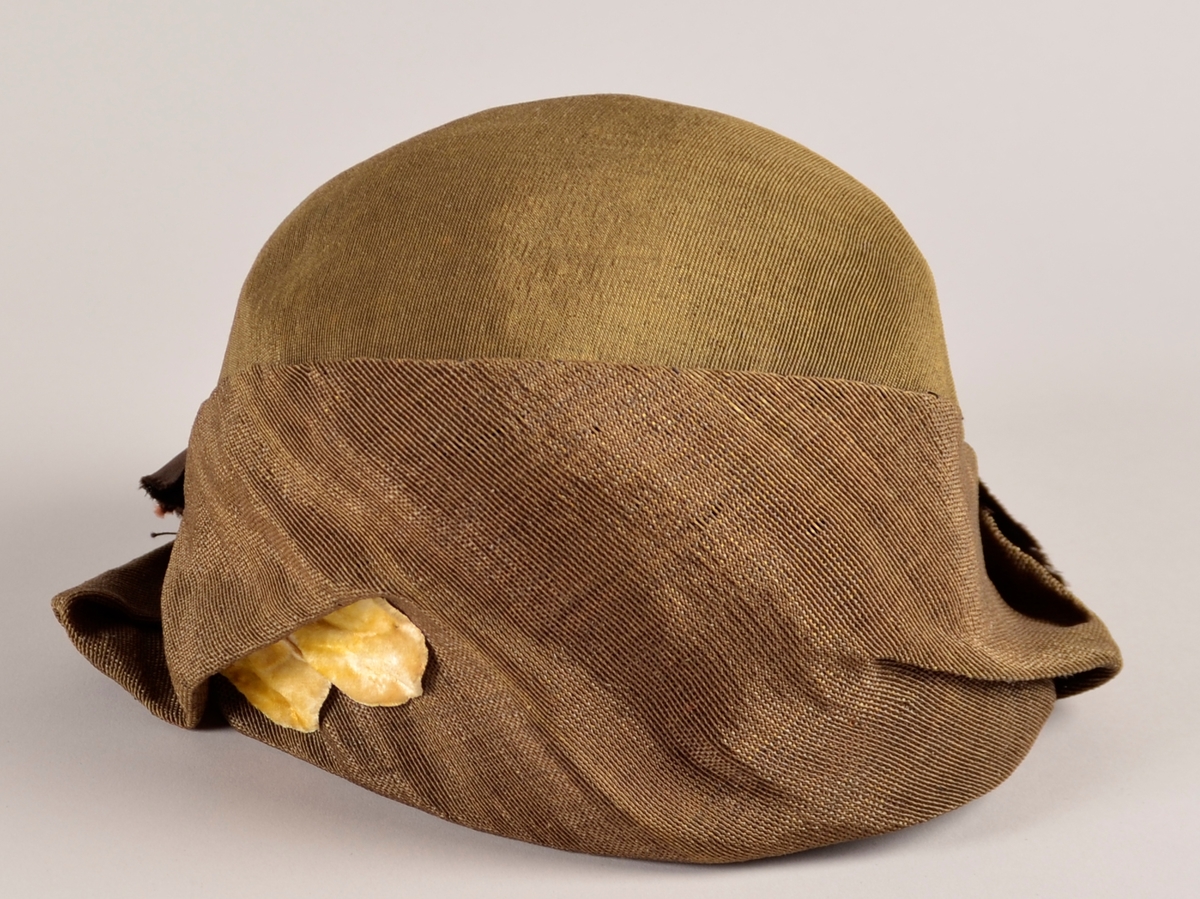 Kvinnehatt. Stiv materiale. Utsida i gyllent, svart/gull, stoff. Sløyfe på sida av brunt band. To gule blad av fløyel til pynt på andre sida. Svart fôr.