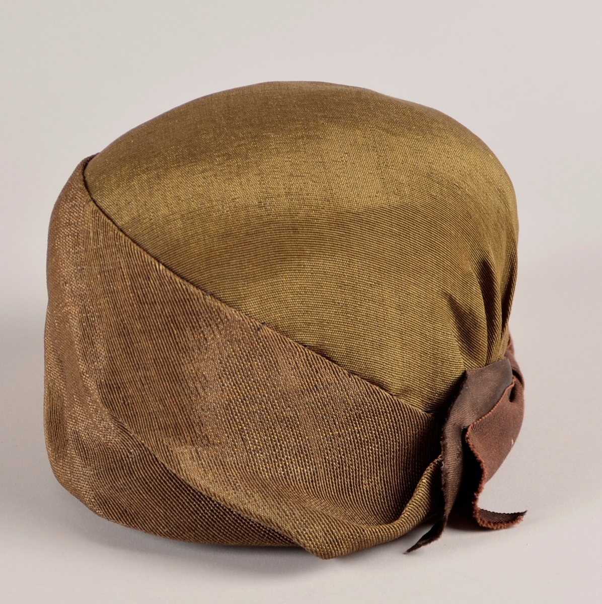 Kvinnehatt. Stiv materiale. Utsida i gyllent, svart/gull, stoff. Sløyfe på sida av brunt band. To gule blad av fløyel til pynt på andre sida. Svart fôr.