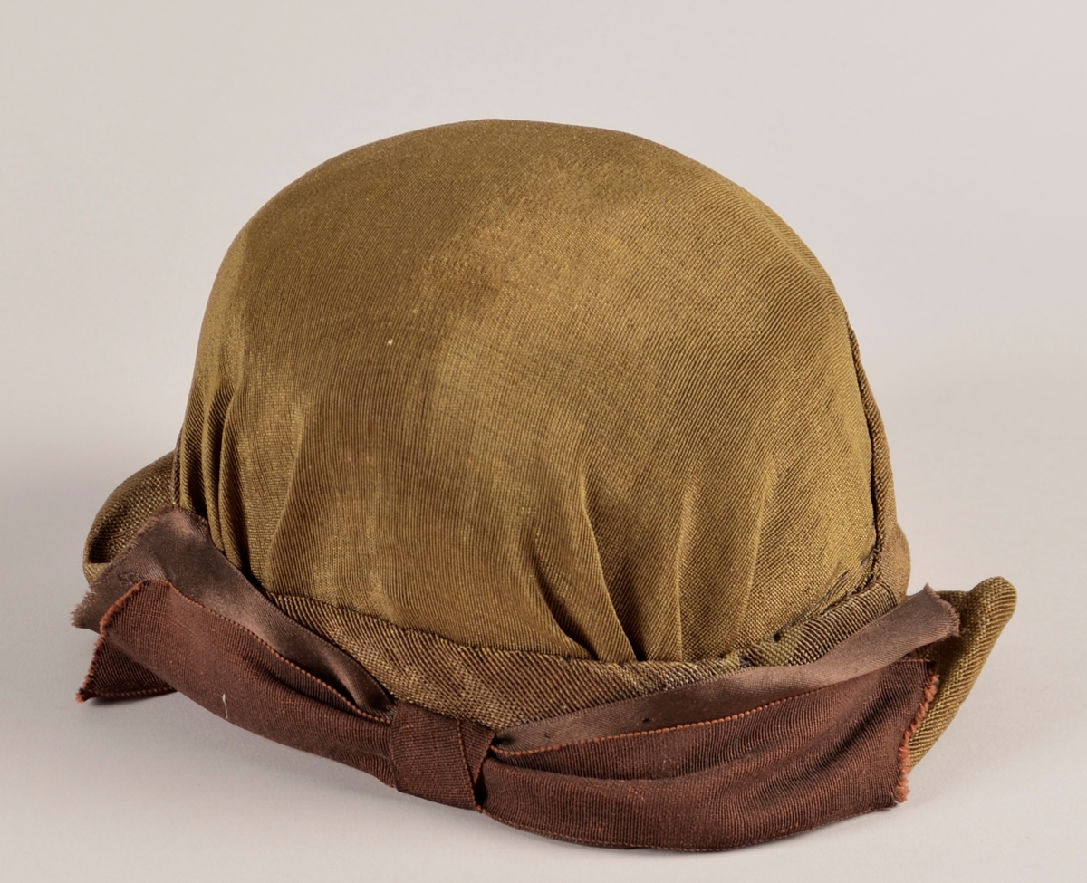 Kvinnehatt. Stiv materiale. Utsida i gyllent, svart/gull, stoff. Sløyfe på sida av brunt band. To gule blad av fløyel til pynt på andre sida. Svart fôr.