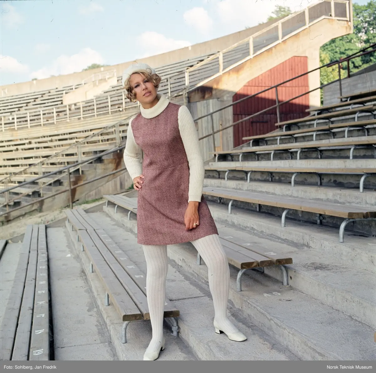 Motebilde, kvinne i lilla ullkjole med hvit genser, sko, strømper og alpelue, på tribunen på idrettsarena. Bildet er en del av en serie der noen er publisert i ukebladet Det Nye 1969, nr. 38.