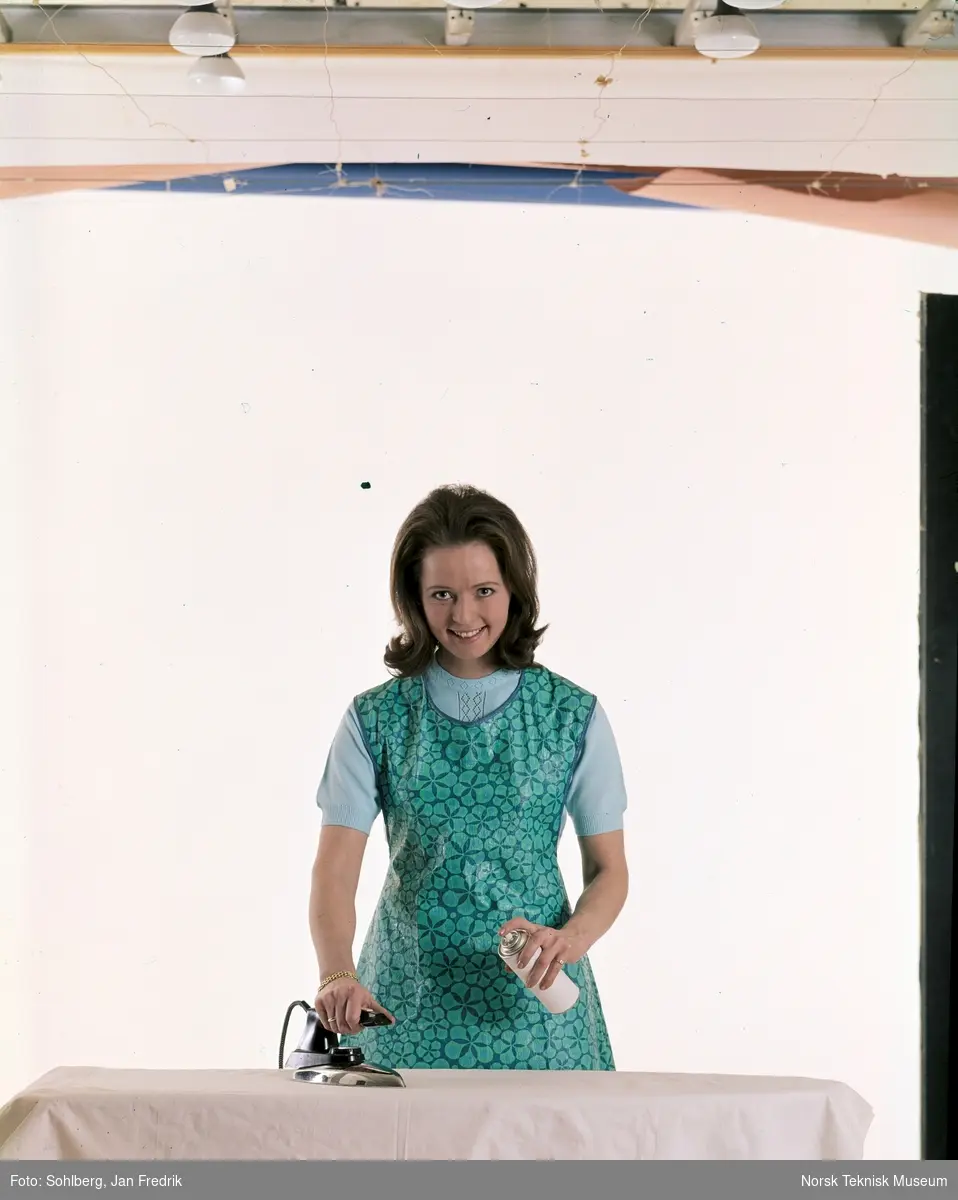 Studiofotografi av en kvinne / husmor som stryker tøy ved et strykebrett. Hun bruker en spray på klærne.