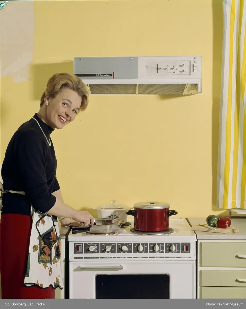 Reklamefoto for komfyr og vifte fra Elektrolux. Med moderne kjøkkenutstyr er livet som husmor ganske enkelt. En smilende kvinnelig modell lager mat ved komfyren.