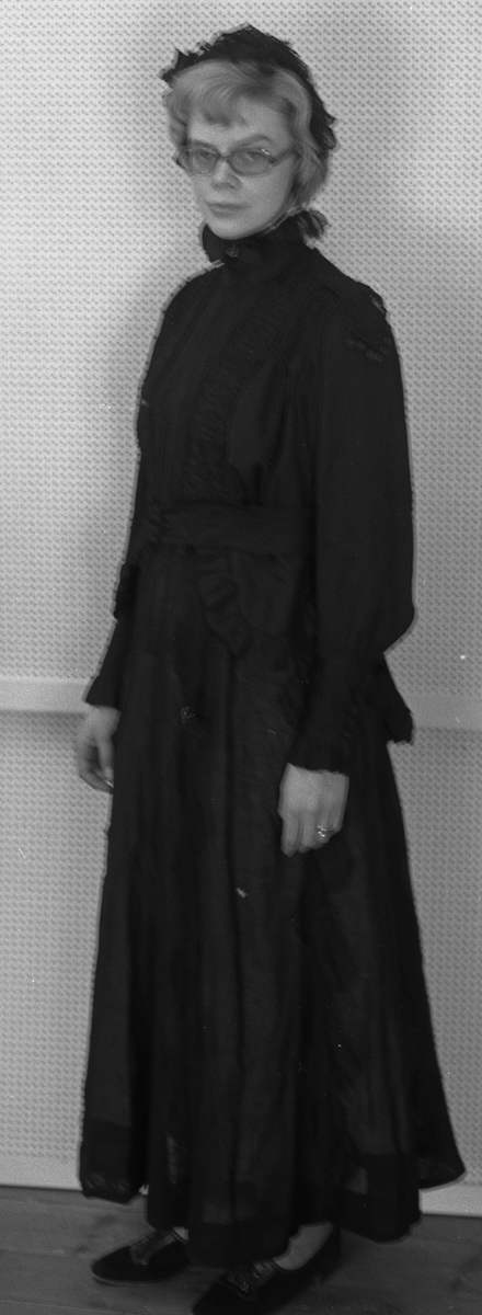 Helforet med svart mugg, 10 fine legg på forstykke som er løst. Bryststykke og halslinning har foldelagt chiffon. Øverst på halslinning rynkekappe av chiffon og smal hvit bobinetteblonde. Brodert chiffonlengde fra halsen og ut over skuldra. Svart blonde ut på skuldrene og på ned i en spiss V foran. Belte i livet. Kappe på hver side under beltet med volang. Kappene fortsetter ned i to snipper bak. Mansjettene på hver erme er høye og lukkes med trykknapper med pynteknapper over.
 