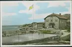 Vykort med motiv från Hönö havsbad med tennisplanen. Två personer står på tennisplanen och utanför havsbadet står två och sitter tre personer, dessa är klädda i 1920-talets mode. Fotografiet är svartvitt men byggnadens tak, gräsytor, havet, himlen och flaggan är kolorerade.