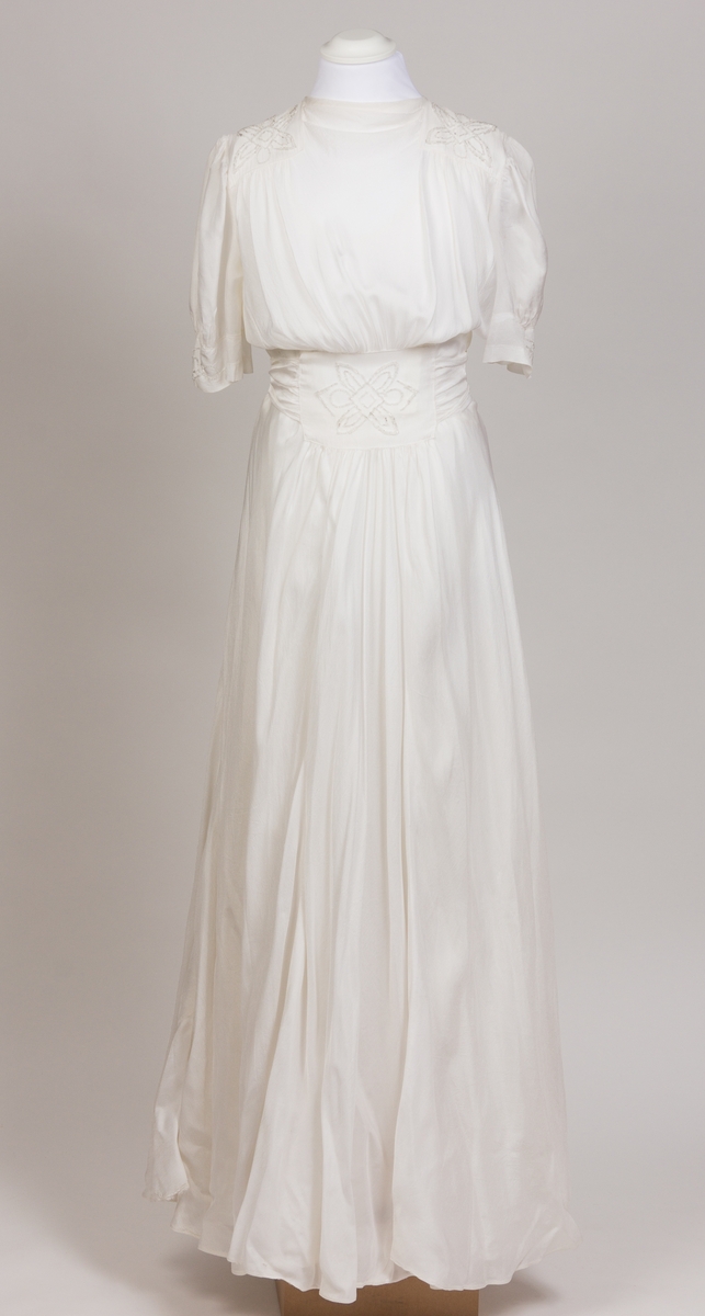 Konfirmasjonskjole, silkekjole dekorert med perlebroderier i front i midje, på skulderpartier og mansjetter. Kjolen er sid med kort erm og lukkes med glidelås og trykkapp i venstre side. Påsydd bredt bånd fra perlebrodert stykke i front som er festet bak med fire knapper.