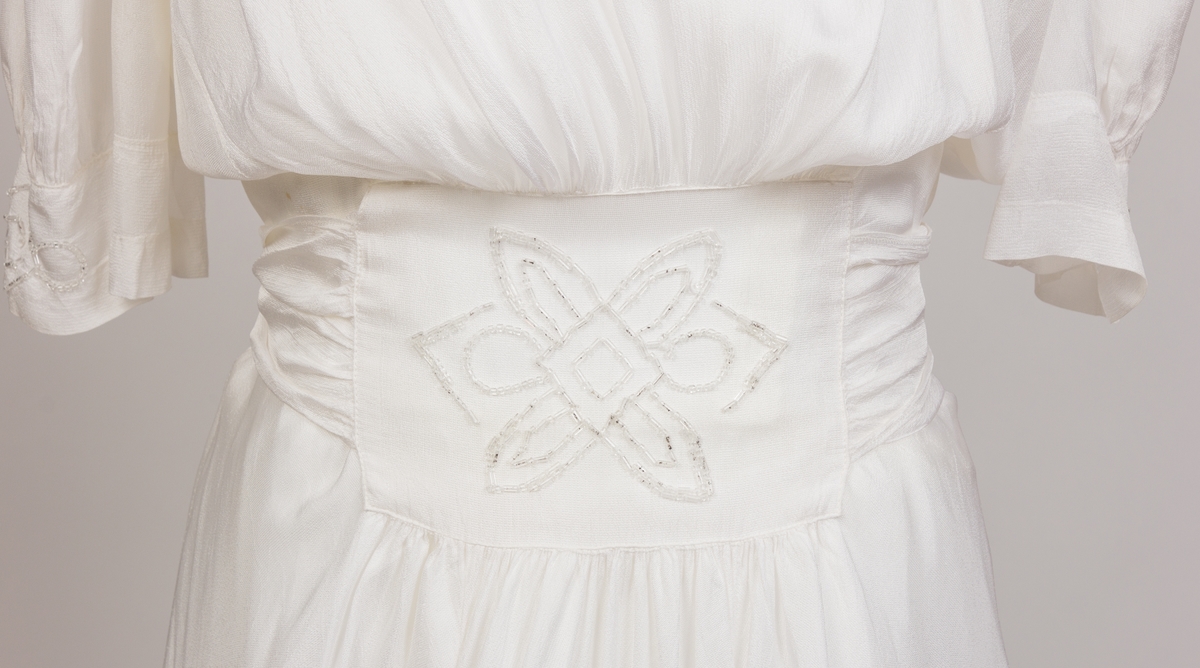 Konfirmasjonskjole, silkekjole dekorert med perlebroderier i front i midje, på skulderpartier og mansjetter. Kjolen er sid med kort erm og lukkes med glidelås og trykkapp i venstre side. Påsydd bredt bånd fra perlebrodert stykke i front som er festet bak med fire knapper.