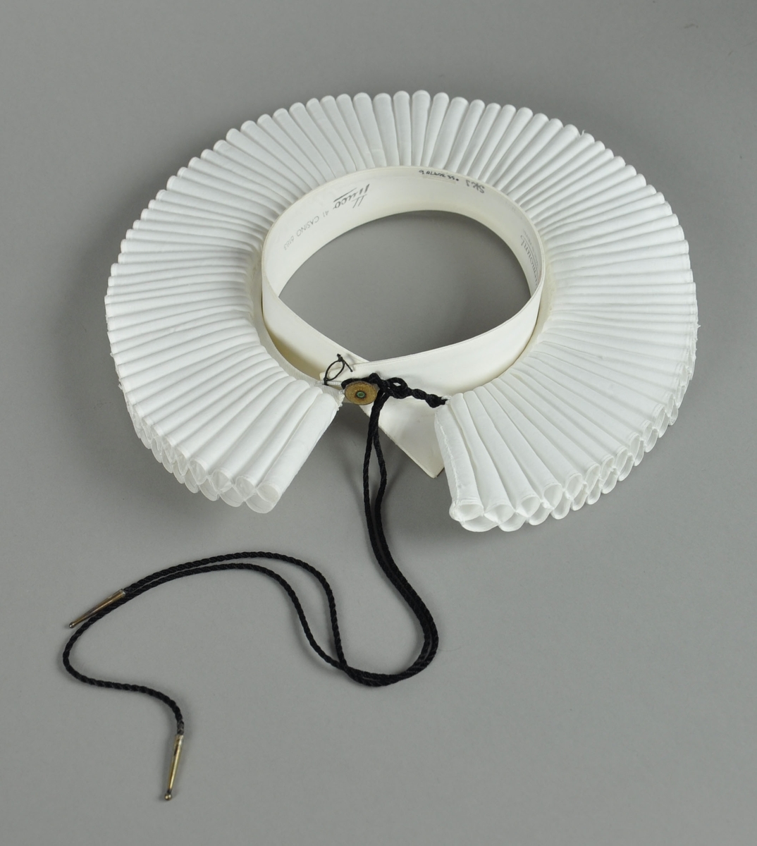 Hvit prestekrage av bomull. Kragen har 2 x 89 piper, 6,5 cm lange. Pipene et sammenfoldet bendelbånd med to hemper og svart messingtupper. Sammen med kragen hører en snipp, også hvit.