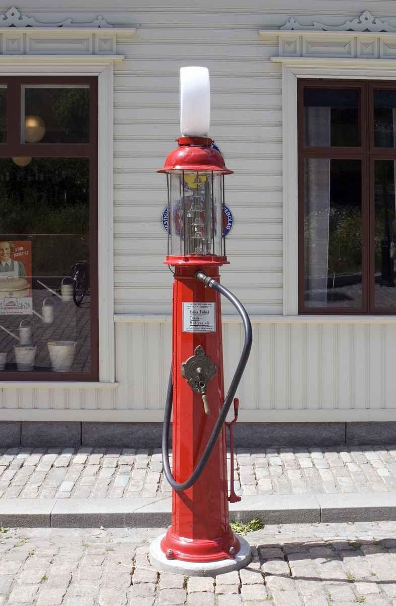 Bensinpump, fabriksgjord. Överst sitter en glasglob, SKAFOR.0015125 på ett metallfäste.  Under globen en cylinder av glas med mätmekanism och gradering. Därunder kommer det rödlackade pumphuset av stål. På en sida sitter en etikett av metall med text: Enligt Kungl. Maj:ts förordning angående eldfarliga oljor är det Förbjudet [understruket] att härstädes Röka Tobak [understruket] samt Tända [understruket] eller Medföra Eld [understruket]. Den som bryter mot sagda föreskrift, äventyrar jämlikt förordningen angående eldfarliga oljor § 60 ansvar av böter från och med 10 t.o.m. 500 kr." Etiketten sitter strax ovanför ett pumphandtag med omgivande dekorativt utformade "bricka" av metall och handtag svarvat av trä. "Brickan" är rund och utformad med graderingar från 0 till 25 med femliters intervaller. Text: PUMP IV-SC 54 A.B. J.C. LJUNGMAN MALMÖ VICI No 6705, ett räkneverk och LITER. Till höger om detta sitter ett annat pumphandtag av rödlackerat stål, fäst nertill. Ovanför etiketten börjar bensinslangen av gummi i ett metallfäste. Den fortsätter i en böj neråt till vänster, runt och upp och avslutas av den utgående kranen av metall, som hänger i ett hål på cylindern. Där bredvid finns också en låsbar lucka.