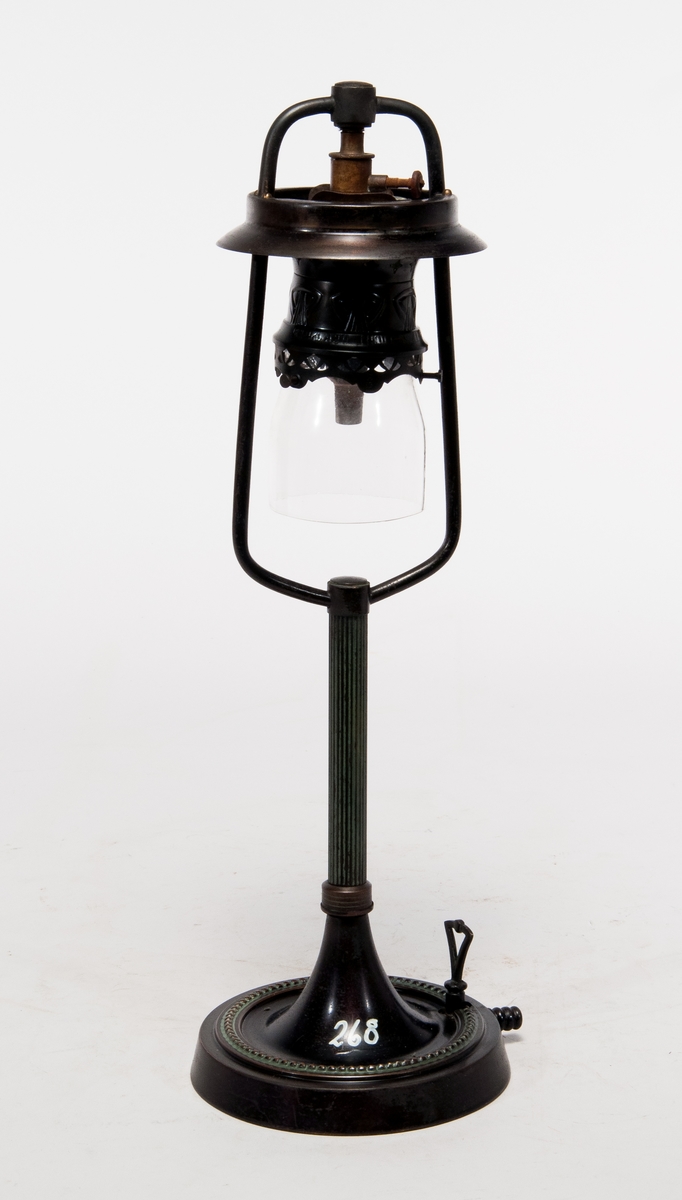 Gaslampa av bordsmodell i metall och glas, märkt: "Graftzin-light D.R.P. 126135". Lös ettikett med text: "Bordstake på svart fot. Bordstakarna härleda sig från 1870-1880 talet, var i bruk intill 1910 talet".
Påmålad märkning "268".