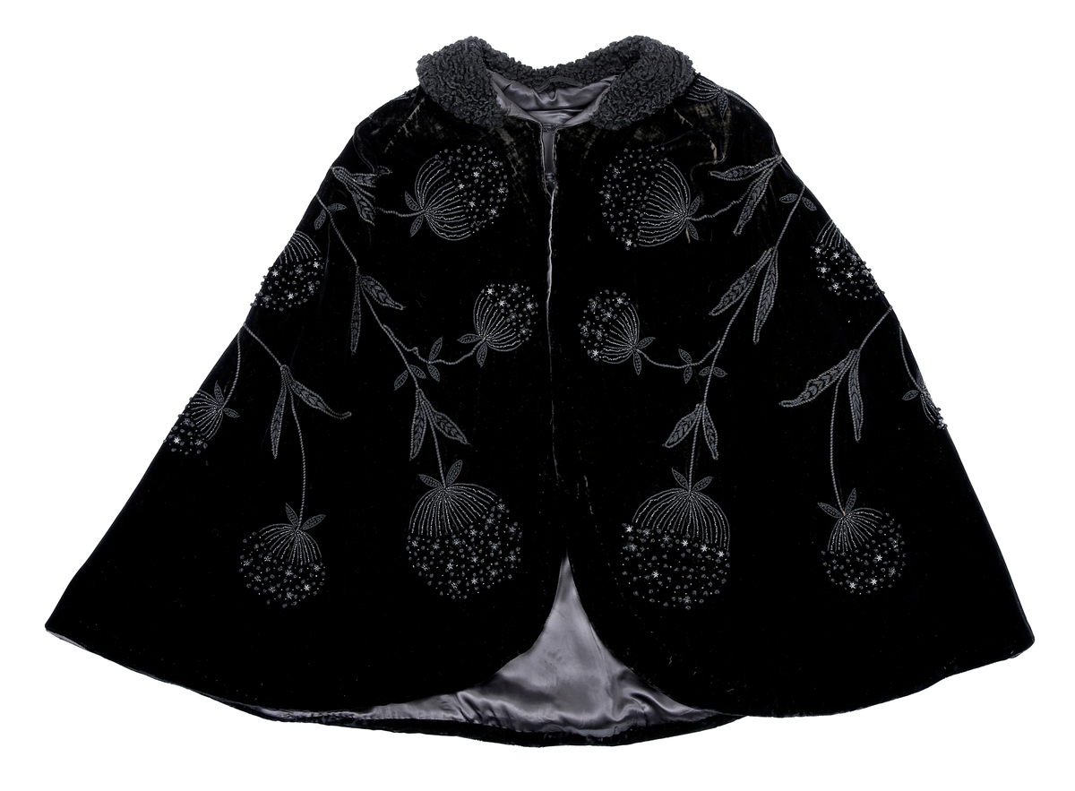 Sort kep i fløyel med lissebroderi, perler og plaststjerner (eller bakelitt?) som former blomster. Krage med persianer. Foret med silke eller kunstsilke. Den har to hemper i halsen. 