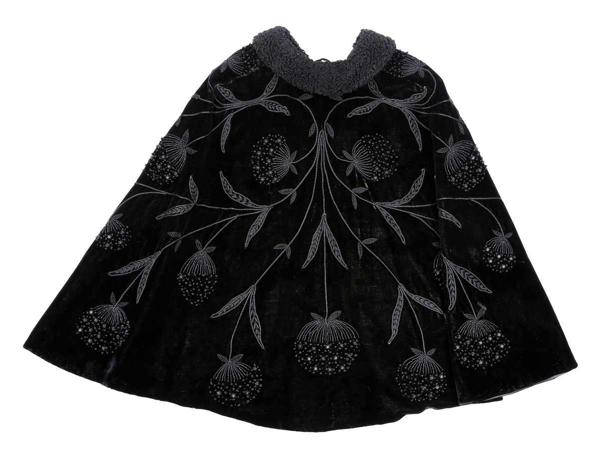 Sort kep i fløyel med lissebroderi, perler og plaststjerner (eller bakelitt?) som former blomster. Krage med persianer. Foret med silke eller kunstsilke. Den har to hemper i halsen. 