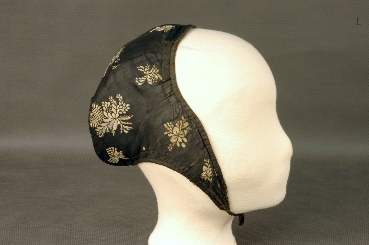 Ørelue i svart silke med hvite broderte roser. Ingen dusk. Foret med lyst linstoff. Svarte knytteband.