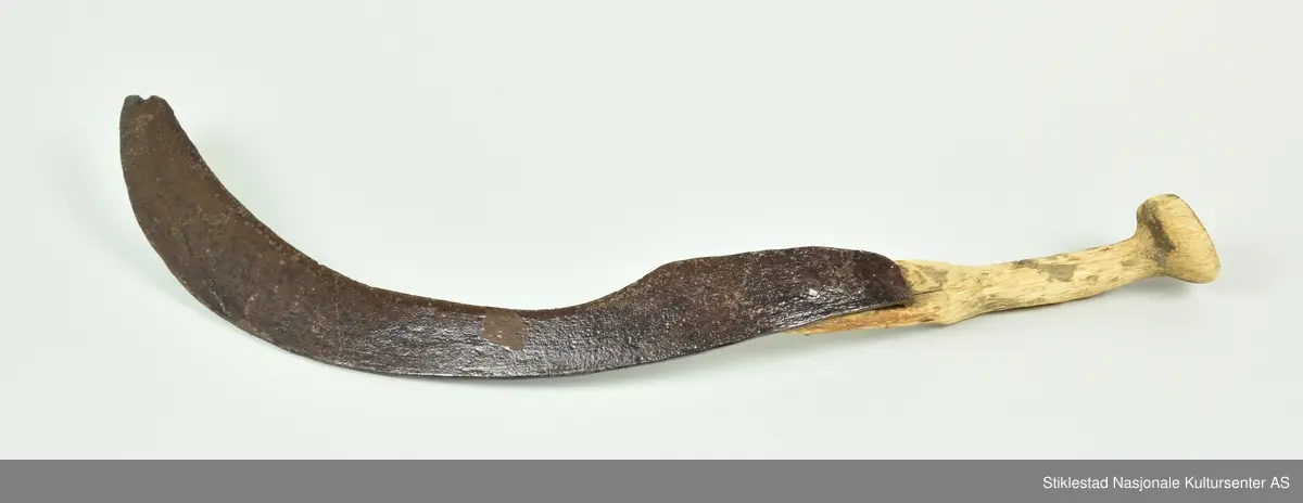 Halvmåneformet blad av jern, festet på ett skaft av naturvokst bjørk (grein). Bladet er festet til skaftet med en jernnagle, samt en tange som går gjennom ett hull i skaftet, er bøyd og banket fast til treet på motsatt side. Skaftet har lett buet form og teljet til for ett godt grep. Noe råteskader på skaftet. Skaftet har halvmåneformet tverrsnitt ved bladfeste. Sigden har ett langt smalt blad som har brukket spiss, samt at skaftet har sprukket. Bladet er merket JL. Bladet er innsatt med Basol, derfor den blanke og svarte overflaten.