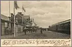 Stationsområde med resande på perrong och vy på stationshus och vagnar.