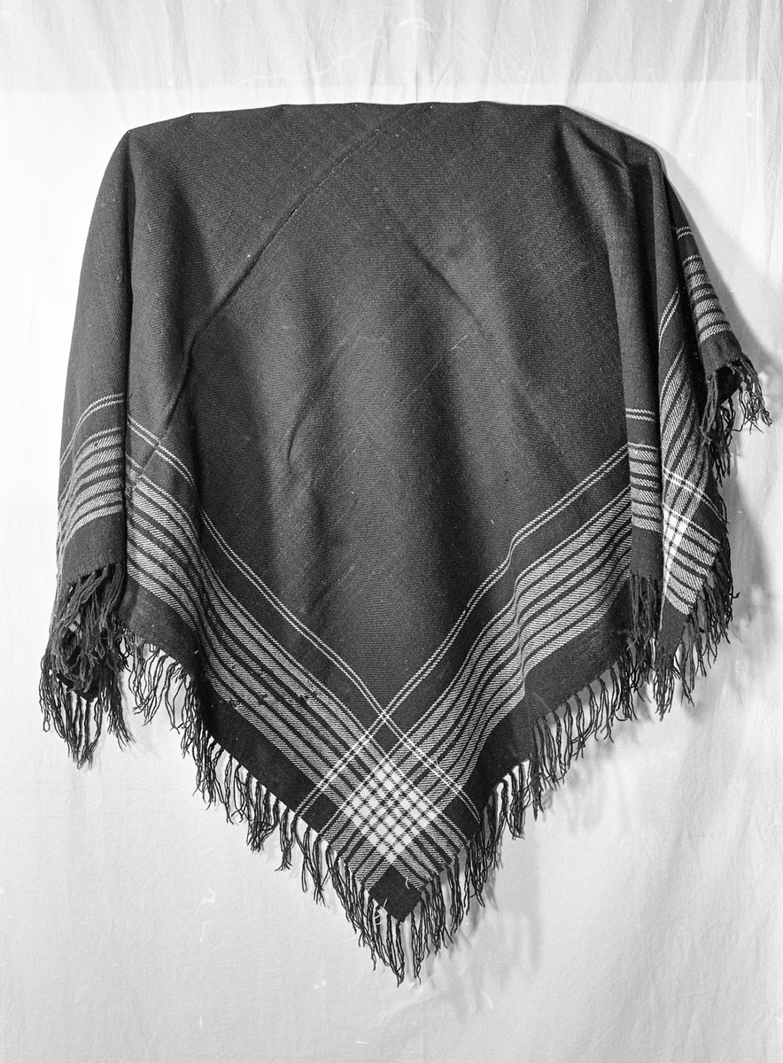 Sjal i ull med frynser på alle kanter. Det er vevd inn mønster bestående av lyse render langs kantene. Det er skjøtt sammen av to stykker, og skjøten er håndsydd og er omtrent på midten. 