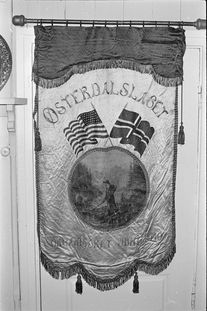 En tømmerfløter i elva, med skog og fjell i bakgrunnen og en solrød himmel. Over dette malte motivet er det amerikanskje og det norske flagget