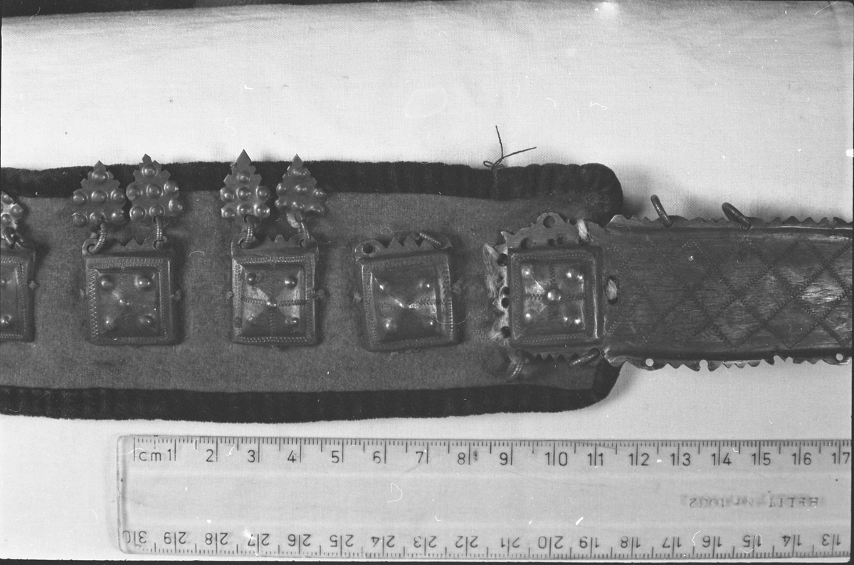 Stølebelte av rødt ullstoff kantet med mørkt ullstoff og med striestoff på innsida. Det er små beltestøler langs hele beltet, som hver har to lauv. Beltespenna har enkel gravert dekor. 