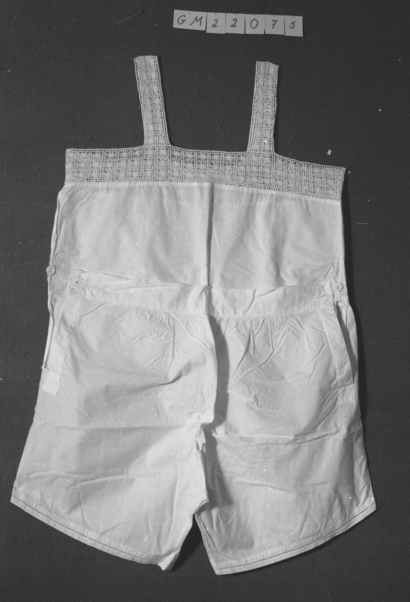 Combination, kvinne. N1b
Ett for-og ett bakstykke, rett oventil, påsydd 6 cm bred heklet kant rundt åpning. Ikke ermer, 3, 5 cm bred heklede skulderstropper. Buksa søm midt foran, 14 l splitt i sidene, kneppet med stofftrukken knapp, søm midt bak, bakstykket til å kneppe ned, 4, 5 cm bred linning, flere små rynkinger på hver side av midtsøm bak. Hullfaldsøm rundt låråpning. Livstykket sprukket i siden fra linning til heklekant.