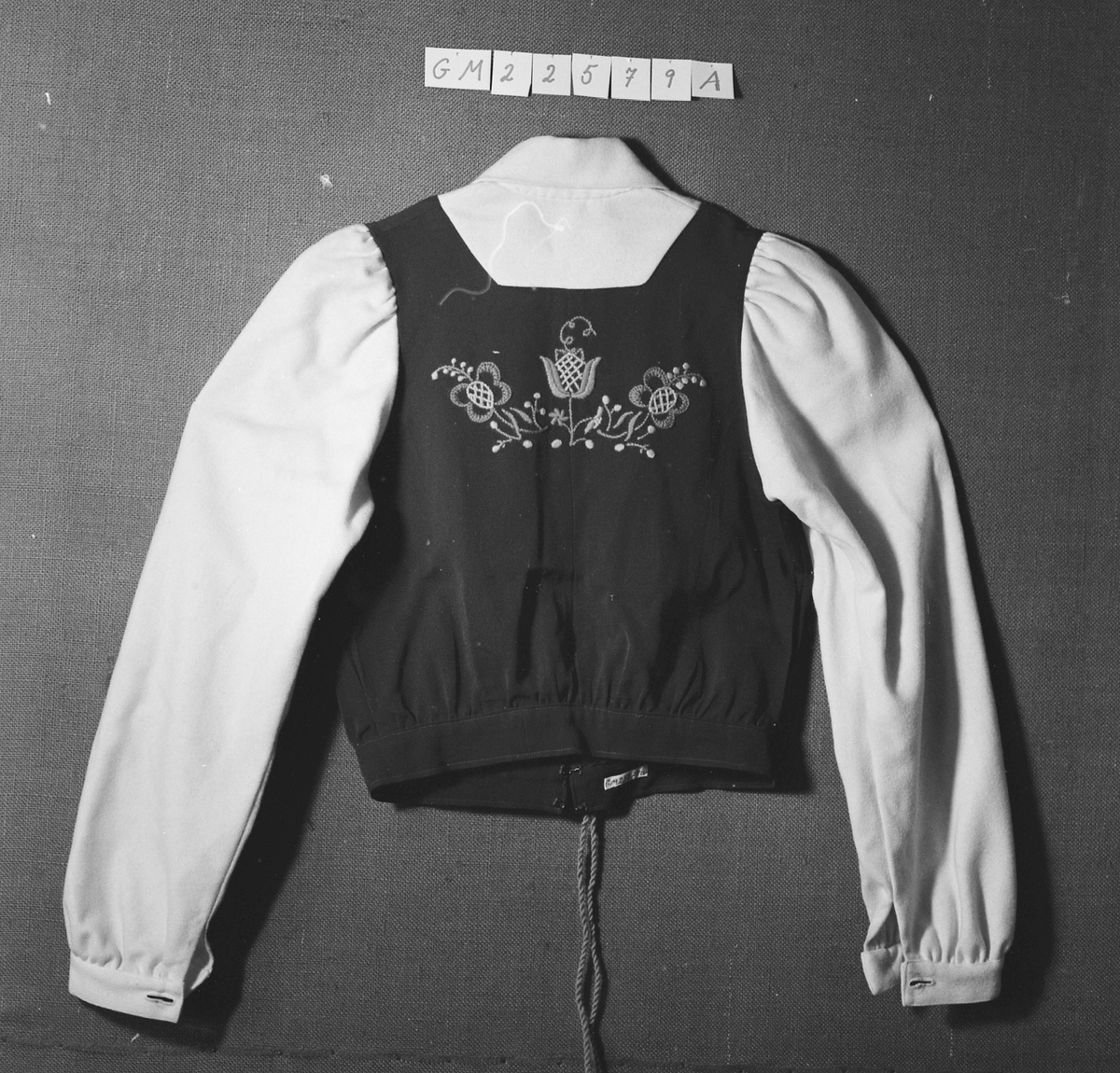 Overdel til drakt bestående av et kypertvevd blått bomullstoff brodert med ullgarn og med ermer og krage av lerretsvevd ull. Front og ermer har tinnknapper med støpt mønster. Det er hekter og trykknapper til å lukke med.
Buksa til drakta er av typen strekkbukse med press og strikk under beina. Den har glidelåslukning i siden og to lommer med glidelås i front.