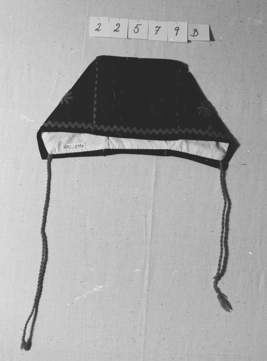 Overdel til drakt bestående av et kypertvevd blått bomullstoff brodert med ullgarn og med ermer og krage av lerretsvevd ull. Front og ermer har tinnknapper med støpt mønster. Det er hekter og trykknapper til å lukke med.
Buksa til drakta er av typen strekkbukse med press og strikk under beina. Den har glidelåslukning i siden og to lommer med glidelås i front.