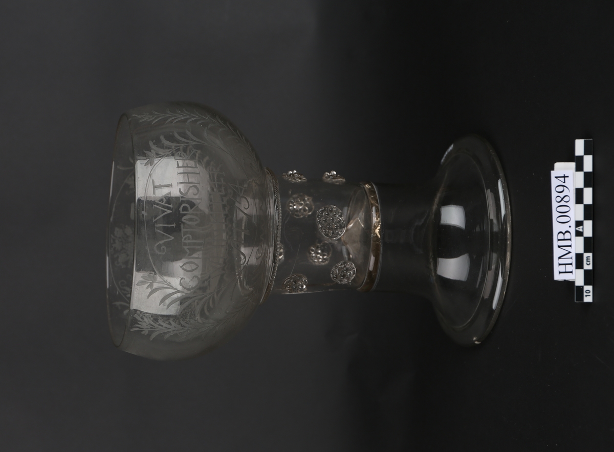 Pokal i glass. Sokkelen er to deler satt sammen oppå hverandre, med en bolle på toppen. Det er inngravert tekst og motiver rundt siden på bollen. Motivet er vinguden Bacchus som sitter på en tønne. Han har vindruer i den ene hånda og en pokal i den andre.