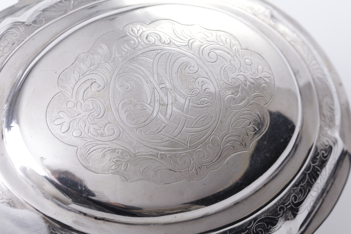 Sockerskrin i silver.
Oval modell. Graverade lambrekäng(?)bårder längs det bukiga livet. Nyckelhålet omges av två blommor i relief (nyckel saknas). Står på fyra inåtvända fötter. På locket ett ornerat spegelmonogram: "AG" (?).  
Stämplar (ofullständiga) + åldermansranka på undersidan av skrinet.
