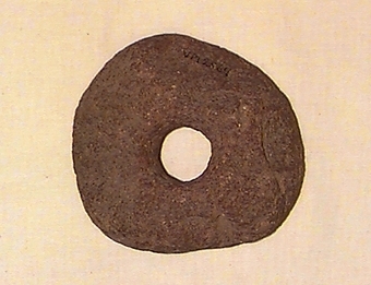 Närmast cirkelrund till formen. Ena sidan plan men kraftigt vittrad, andra sidan "urgrävd" utåt kanterna, högst invid det centralt placerade hålet.