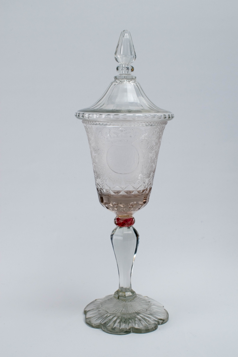 Pokal med lokk i klart glass. Konisk kupa, hvis ytterside er dekorert med sliping og gravering. På forsiden en fremstilling av Juliane Maries kronede monogram med initalene "J M", omgitt av skjell, blomster og bladverk. Balusterformet stett med fasettering, hvis overgang til kupa er markert med en flattrykket rødfarget vulst. Åttekantet fasettert fotplate.