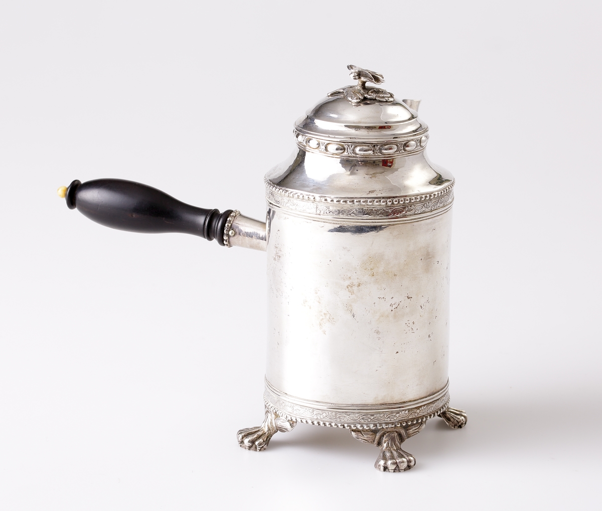 Kaffekanna i silver. 
Cylindrisk modell på tre tassfötter. Graverad bård nedtill, följd av pärlbård. Välvt lock med graverad bård, pärlbård, äggstavsbård och knopp i form av en ros. Försedd med svarvat, svärtat trähandtag, med benknopp. 
Stämplar på undersidan av kannan.