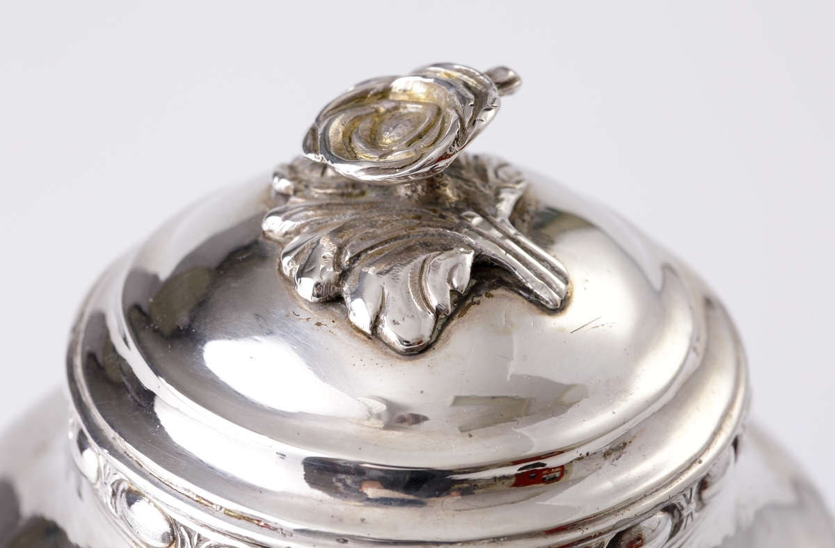 Kaffekanna i silver. 
Cylindrisk modell på tre tassfötter. Graverad bård nedtill, följd av pärlbård. Välvt lock med graverad bård, pärlbård, äggstavsbård och knopp i form av en ros. Försedd med svarvat, svärtat trähandtag, med benknopp. 
Stämplar på undersidan av kannan.