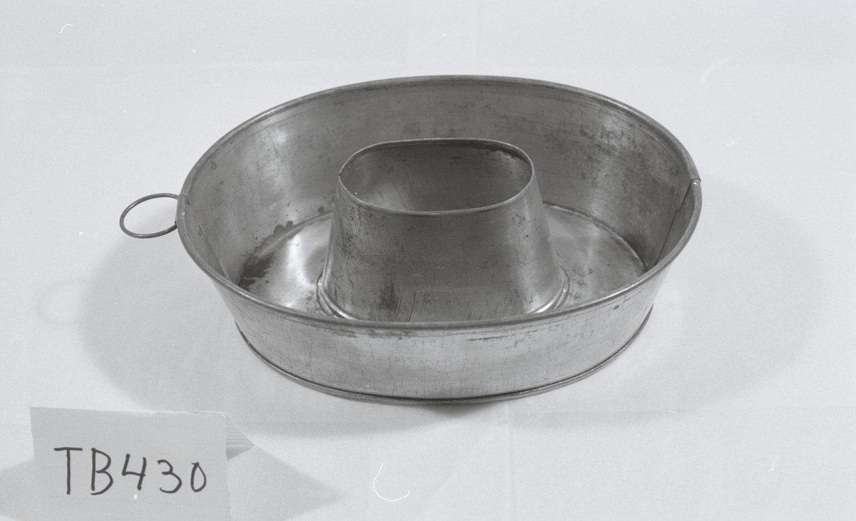 Den ovale puddingformen er svakt konisk og har flat bunn med et ovelt hull med mål B: 5 cm og L: 8 cm. 
Produktet er et bikksmedarbeid.