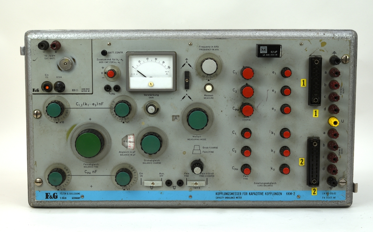 Felten & Guilleaume Kopplungmesser für Kapazitive Kopplungen KKM-2. Mätinstrument som använts för att mäta och eliminera kapacitiv koppling (:1). Höljet av plåt är målat med grå hammarlack. Det är rektangulärt och har rundade kanter och hörn samt ett avtagbart lock av plåt som fästs med fyra spännen (:2). Manöverpanelen är av eloxerad aluminiumplåt som även är målad i en grågrön nyans med text på tyska och engelska tryckt i svart. På panelens nederkant finns en blå rand med tillverkarlogotyp, modellnamn och artikelnummer tryckt i svart. I det övre vänstra hörnet finns en separat panel med banankontakter för extern strömförsörjning och strömbrytare och under panelen finns plats för nio batterier av R20-typ.

För inställningar och justeringar finns en mängd reglage i form av vred och skjutreglar. Vreden har rattar i olika storlek av svart plast med olika färg på topparna för att indikera funktionen. På den vänstra delen av panelen finns sju gröna rattar i olika storlek, tre mindre rattar med grå topp, en röd ratt och tre skjutreglar. Där finns också en rektangulär amperemätare med vit mätartavla och grå ram tillverkad av Gossen. Den största gröna ratten "Feinabgleich" och den lite mindre ratten "Grobabgleich" har ett gemensamt fönster där vita skalskivor med svart och röd text syns. Även ratten "Meßart" har ett skalfönster.

På den högra delen av panelen finns två rader med sex röda rattar vardera följt av två avlånga multipinkontakter i svart plast och en rad med elva banankontakter med bruna och svarta höljen samt en med gult hölje. Ovanför raderna med röda rattar finns en avtagbar kondensator i bakelit som sitter i banankontakter.

På höljets undersida finns två trämedar och på kortsidorna finns utfällbara metallhandtag. Bakom handtagen finns spår av någon form av märkning i tusch som nötts bort, endast "Cst" är läsligt (dvs Stockholm C). På locket finns spår av gula etiketter, möjligen adresslappar. På den högra kortsidan finns en plakett med tillverkarnamn, modellnamn och artikelnummer. På denna finns rester av etiketter.

Till apparaten hör två mätkablar (:3-4) som ansluts till multipinkontakterna. På kabeln finns ett handtag med trycke och i änden fyra hål med kontakter som bryts med trycket. På ovansidan av handtaget finns en etikett med tillverkarlogotyp och produktnamn, "Anlegezange AZ-4" med artikelnummer. Där finns även fyra försänkningar i gult, rött, grönt och blått som identifierar kontakterna på änden. Under logotypetiketten finns en röd LED-lampa. Kabelns kontakt har ett hölje av svartmålad metall.