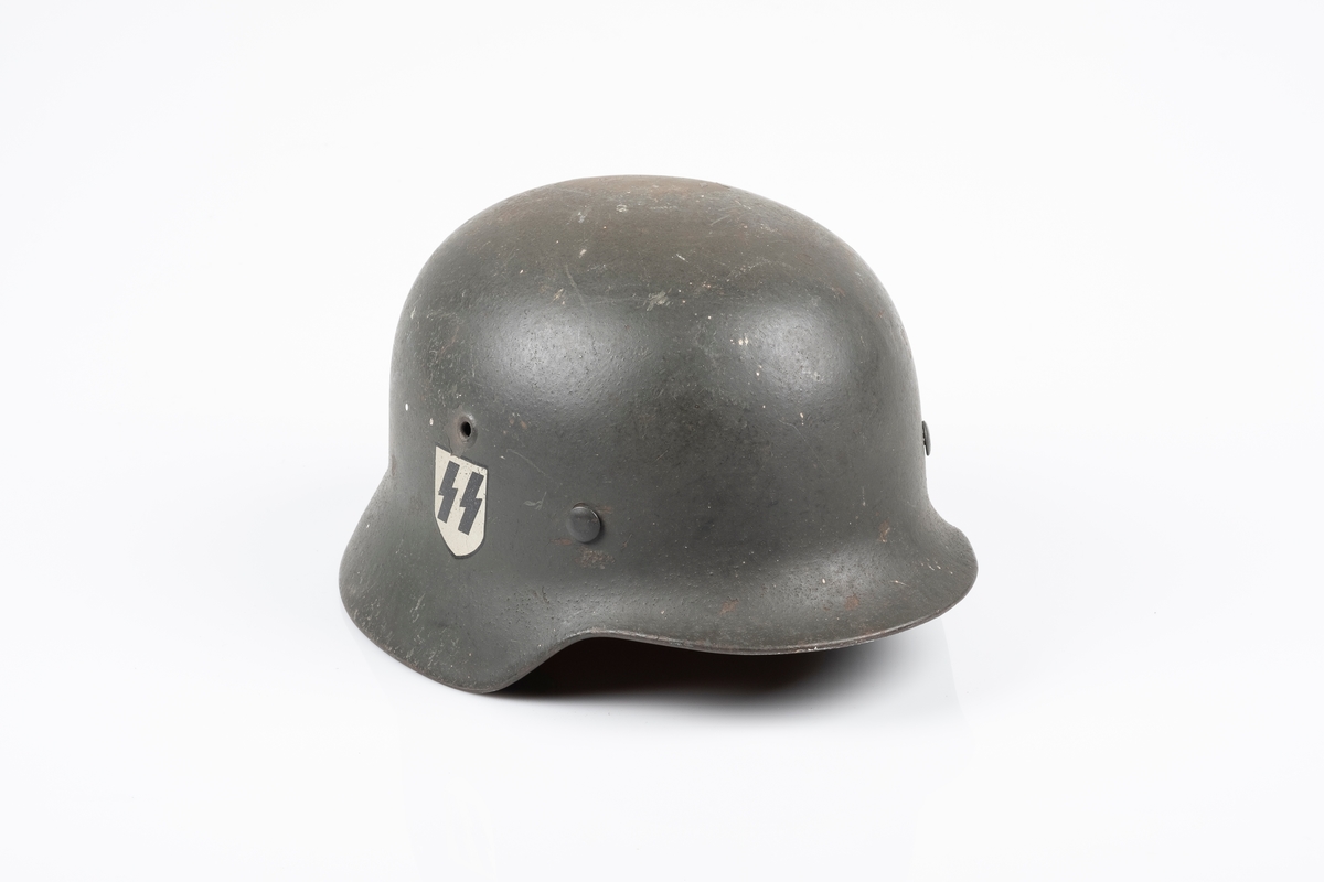 Tysk soldathjelm i metall med "SS" logo. Hjelmen har to sirkelrunde hull på hver side, som er symmetrisk plassert i forhold til hverandre. På innsiden er det festet en innerhjelm av lær med snor som brukes til å tilpasse hjelmen etter hodets størrelse. Hjelmen har lærrem med metallspenne. Hjelmen er malt brun-grønn.